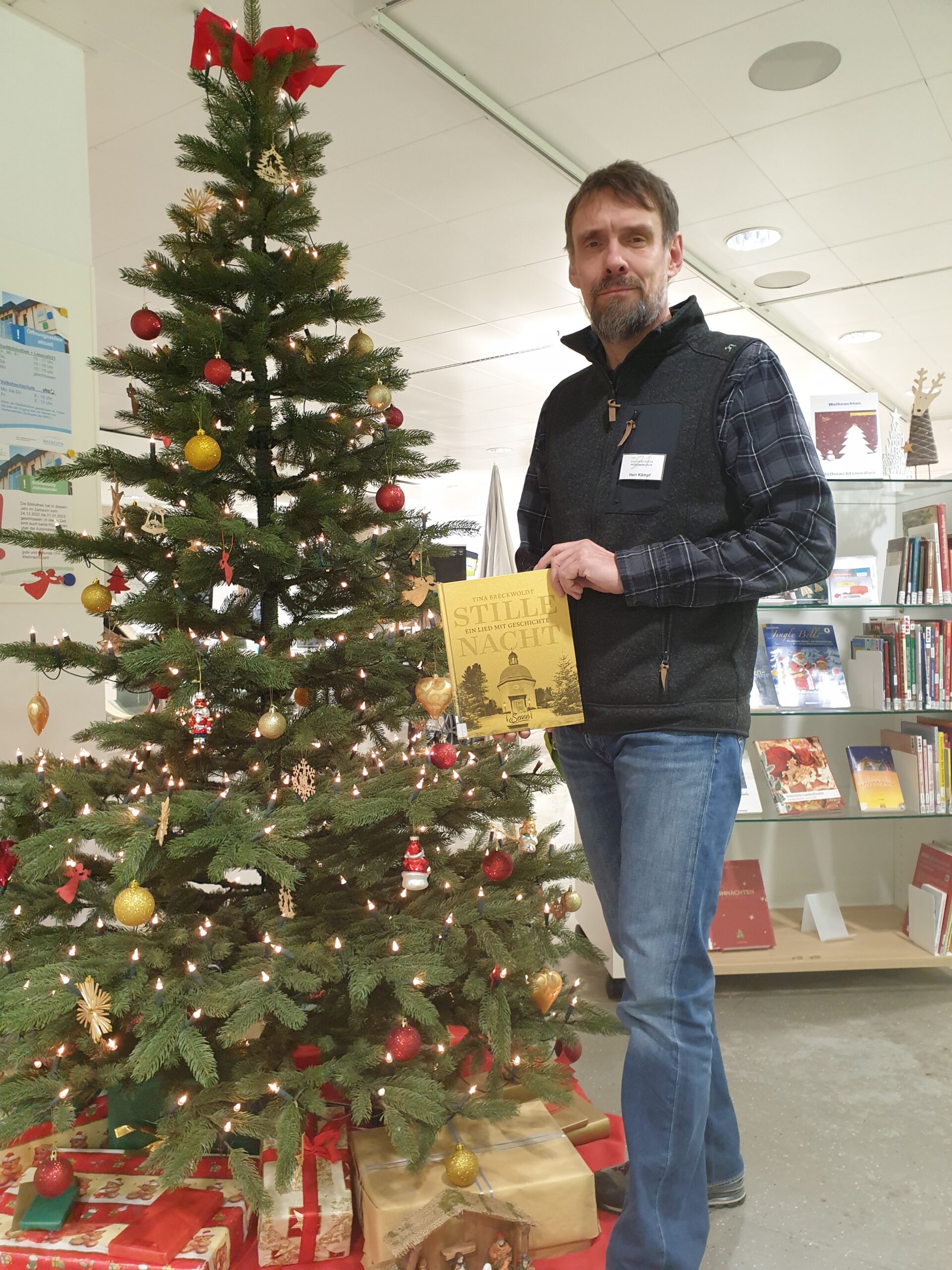 Bibliotheksmitarbeiter steht vor geschmücktem Weihnachtsbaum und hält ein Buch in den Händen.
