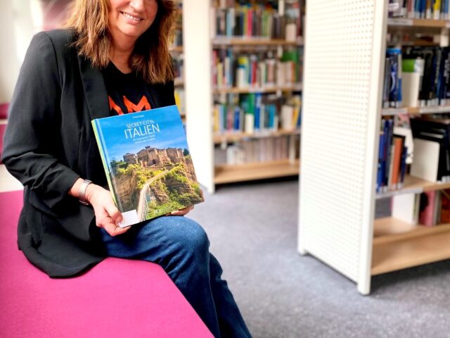 Frau sitzt auf einer Bank in der Bibliothek und hält ein Buch über Italien in die Kamera
