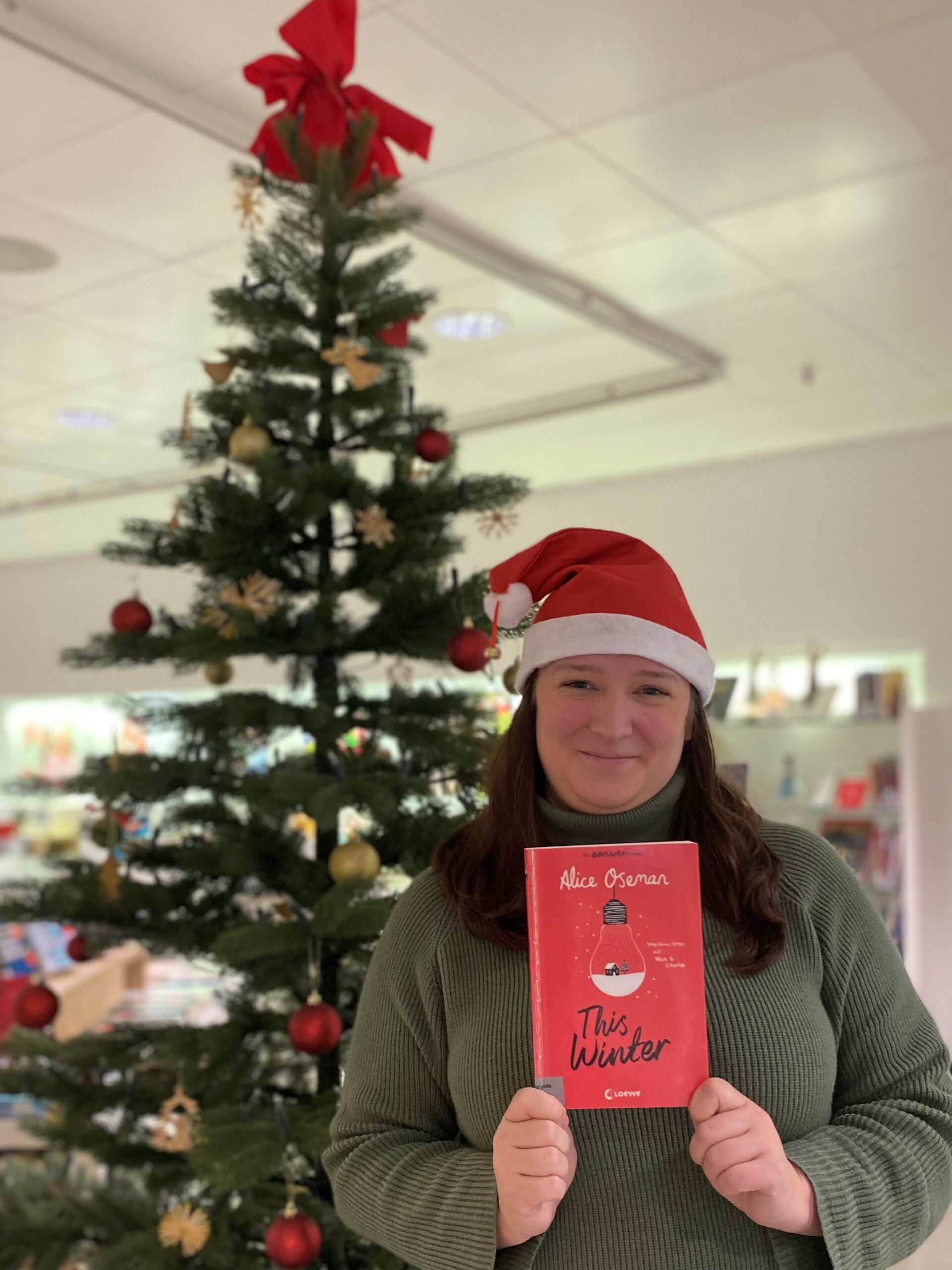 Frau mit Nikolausmütze steht vor einem geschmückten Weihnachtsbaum und hält ein Buch hoch.