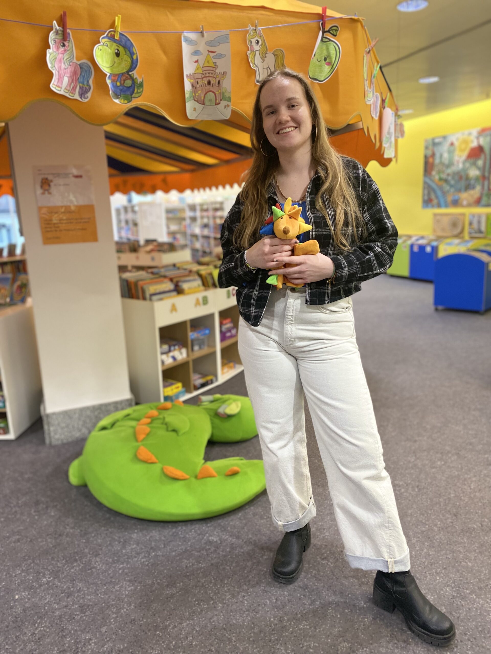 Junge Frau steht in einer Kinderbibliothek mit Stoffigel in der Hand.