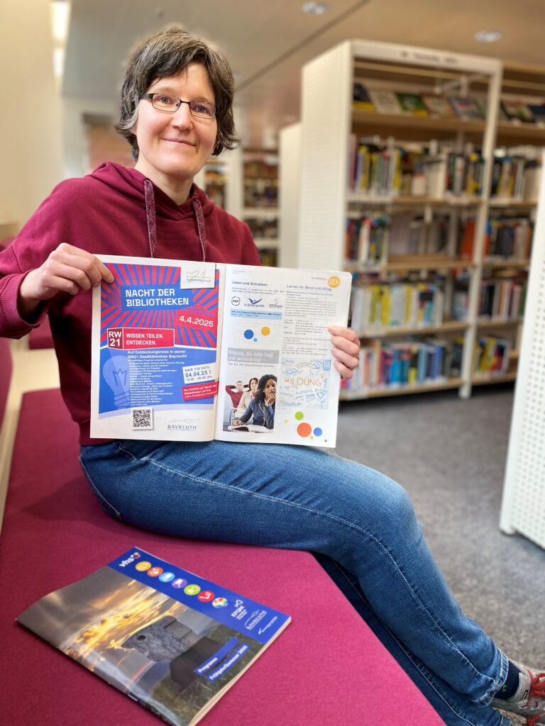 Frau hält das neue Programmheft der vhs und sitzt in einer Bibliothek.