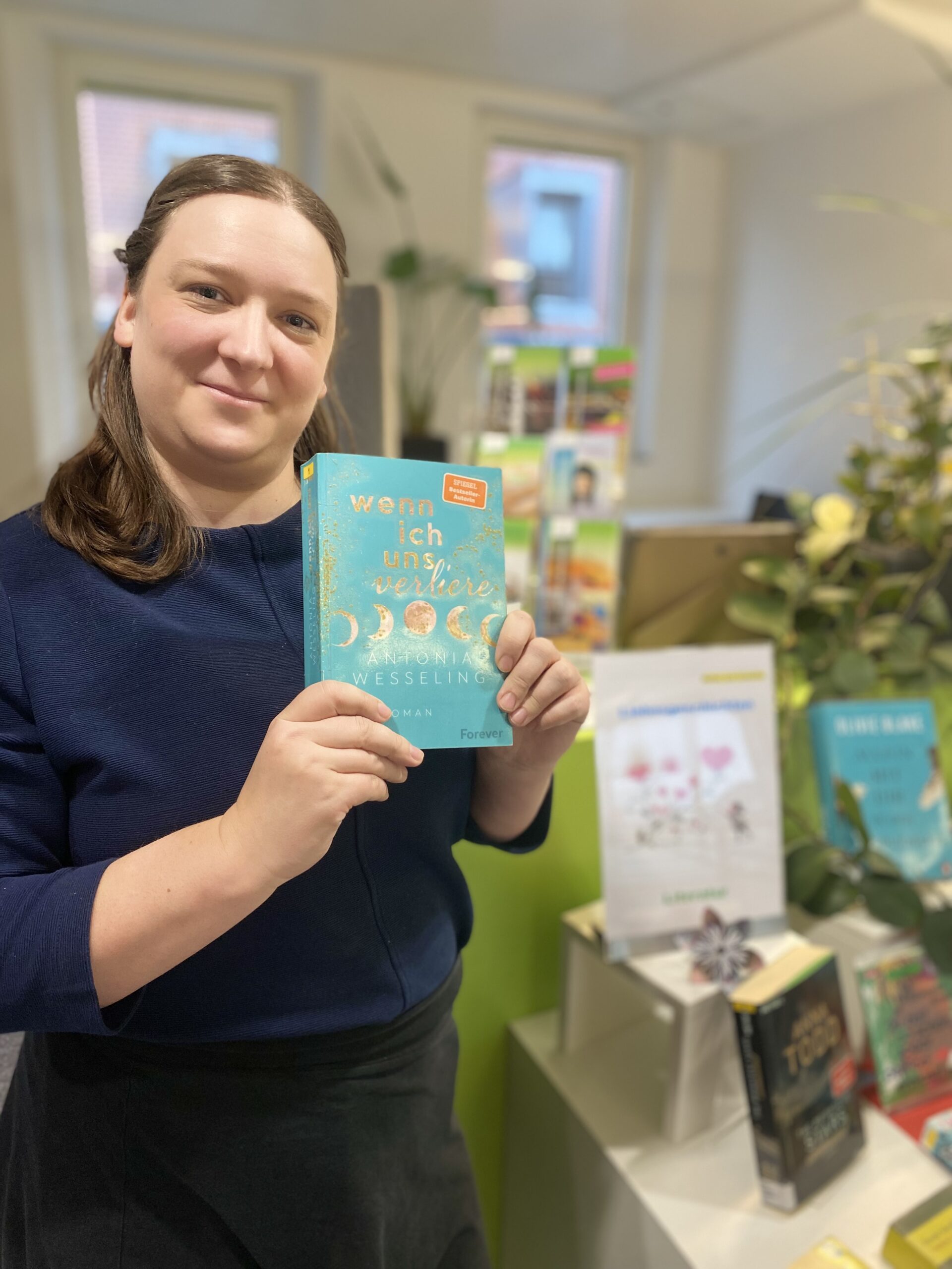 Frau mit blauem Pullover hält einen Roman in die Kamera