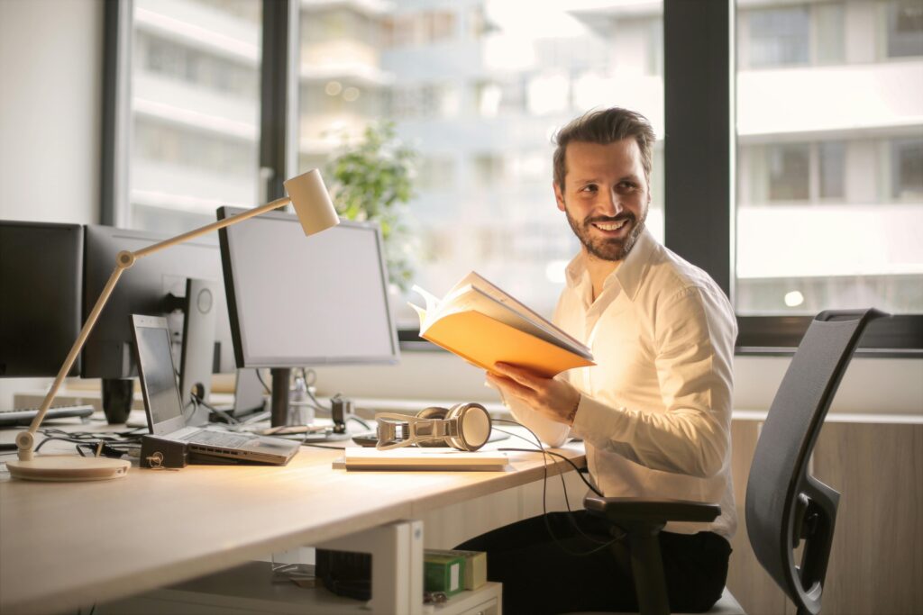 Ein Mann sitzt am Schreibtisch mit einem Buch in der Hand und lächelt. Vor ihm auf dem Schreibtisch stehen Laptop, Bildschirm, Lampe und Kopfhörer.