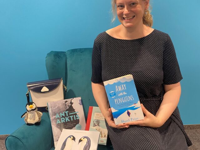 Eine blonde Frau sitzt vor einem blauen Hintergrund auf der Lehne eines blauen Sessels und hält ein Buch lächelnd in die Kamera. Auf dem Sessel sind weitere Bücher, ein Pinguinplüschtier und eine Tasche.
