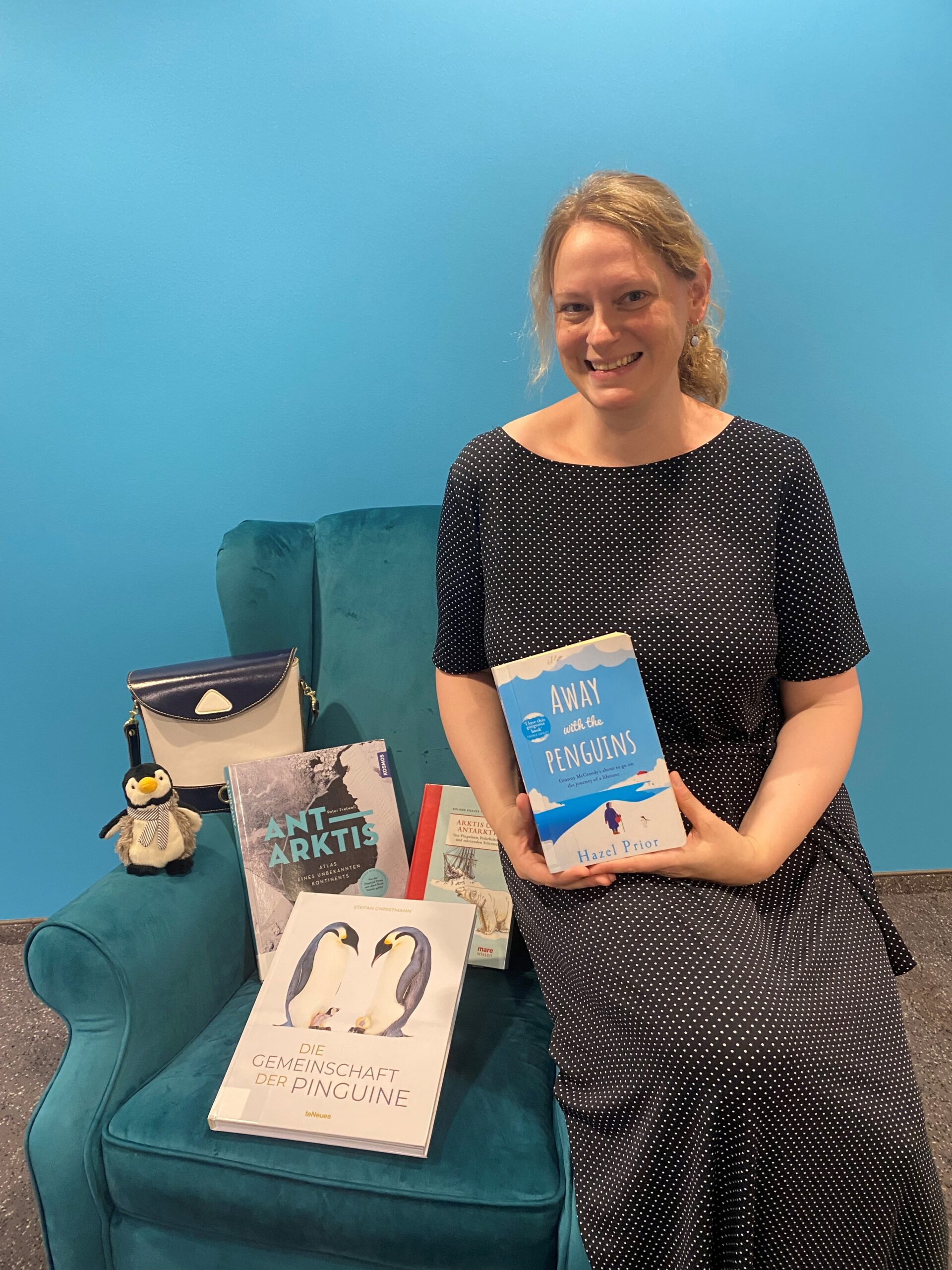 Eine blonde Frau sitzt vor einem blauen Hintergrund auf der Lehne eines blauen Sessels und hält ein Buch lächelnd in die Kamera. Auf dem Sessel sind weitere Bücher, ein Pinguinplüschtier und eine Tasche.