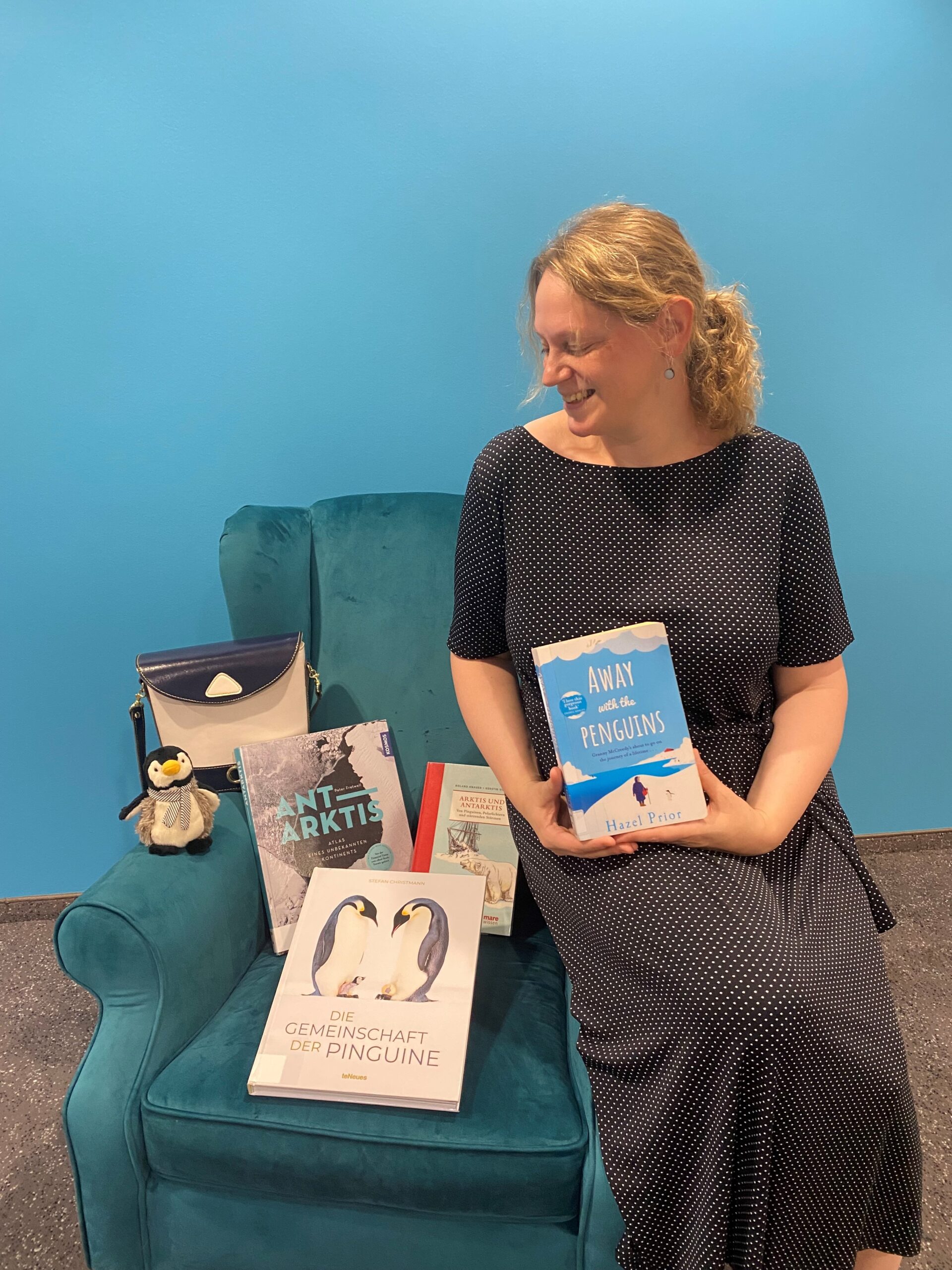 Eine blonde Frau sitzt vor einem blauen Hintergrund auf der Lehne eines blauen Sessels und hält ein Buch in der Hand und schaut lächelnd zum Sessel herunter. Auf dem Sessel sind weitere Bücher, ein Pinguinplüschtier und eine Tasche.