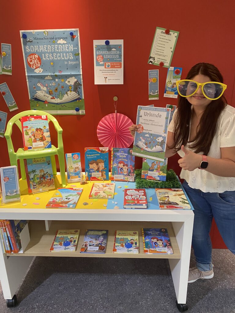 Ein Tisch voller Bücher und Dekorationen, daneben eine junge Frau mit langen braunen Haaren und einer übergroßen gelben Brille, die Daumen hoch in die Kamera zeigt.