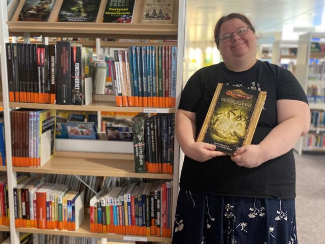 Eine junge Frau mit schwarzer Kleidung und Brille hält lächelnd ein Buch in die Kamera. Neben ihr ist ein Regal mit Büchern.