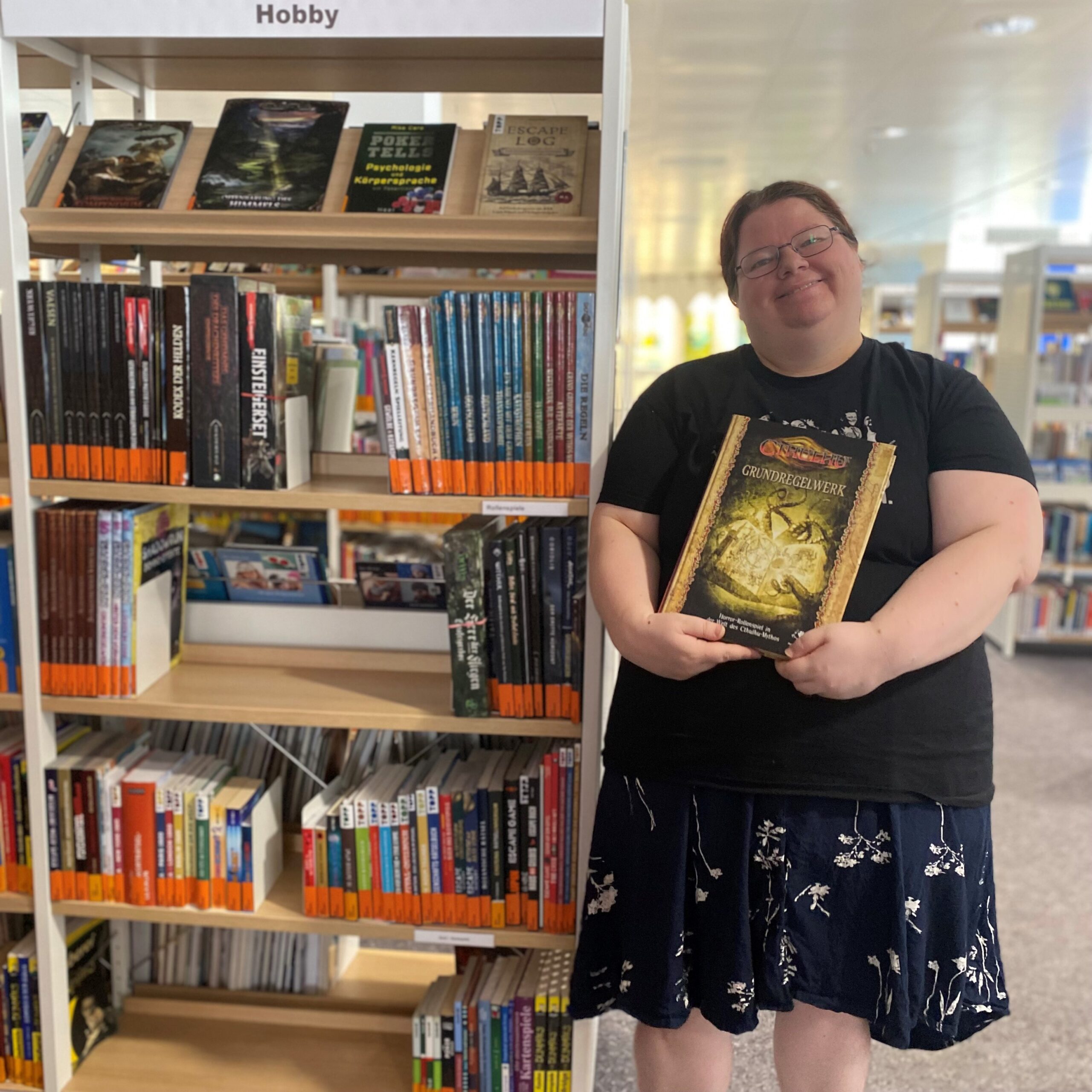 Eine junge Frau mit schwarzer Kleidung und Brille hält lächelnd ein Buch in die Kamera. Neben ihr ist ein Regal mit Büchern.