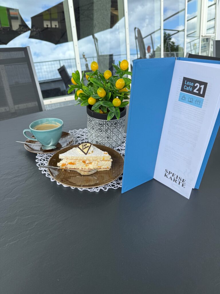 Eine volle Kaffeetasse, ein Teller mit einem Stück Kuchen und eine Preiskarte stehen auf einem Tisch draußen. Im Hintergrund ist eine Zitronenblume zu sehen.