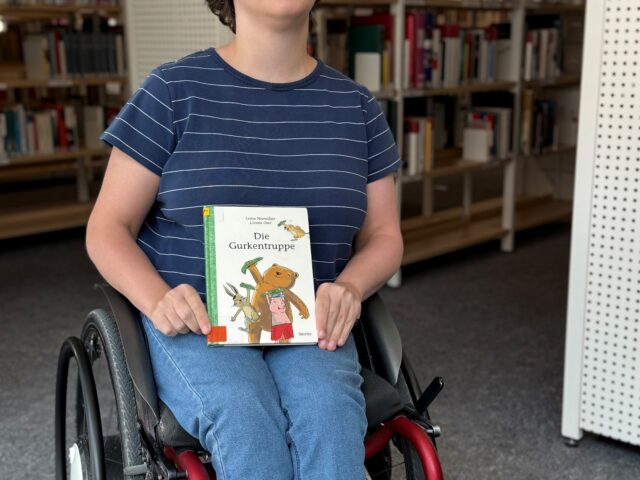 Eine junge Frau im Rollstuhl, die ein Kinderbuch in der Hand hält und in die Kamera lächelt.