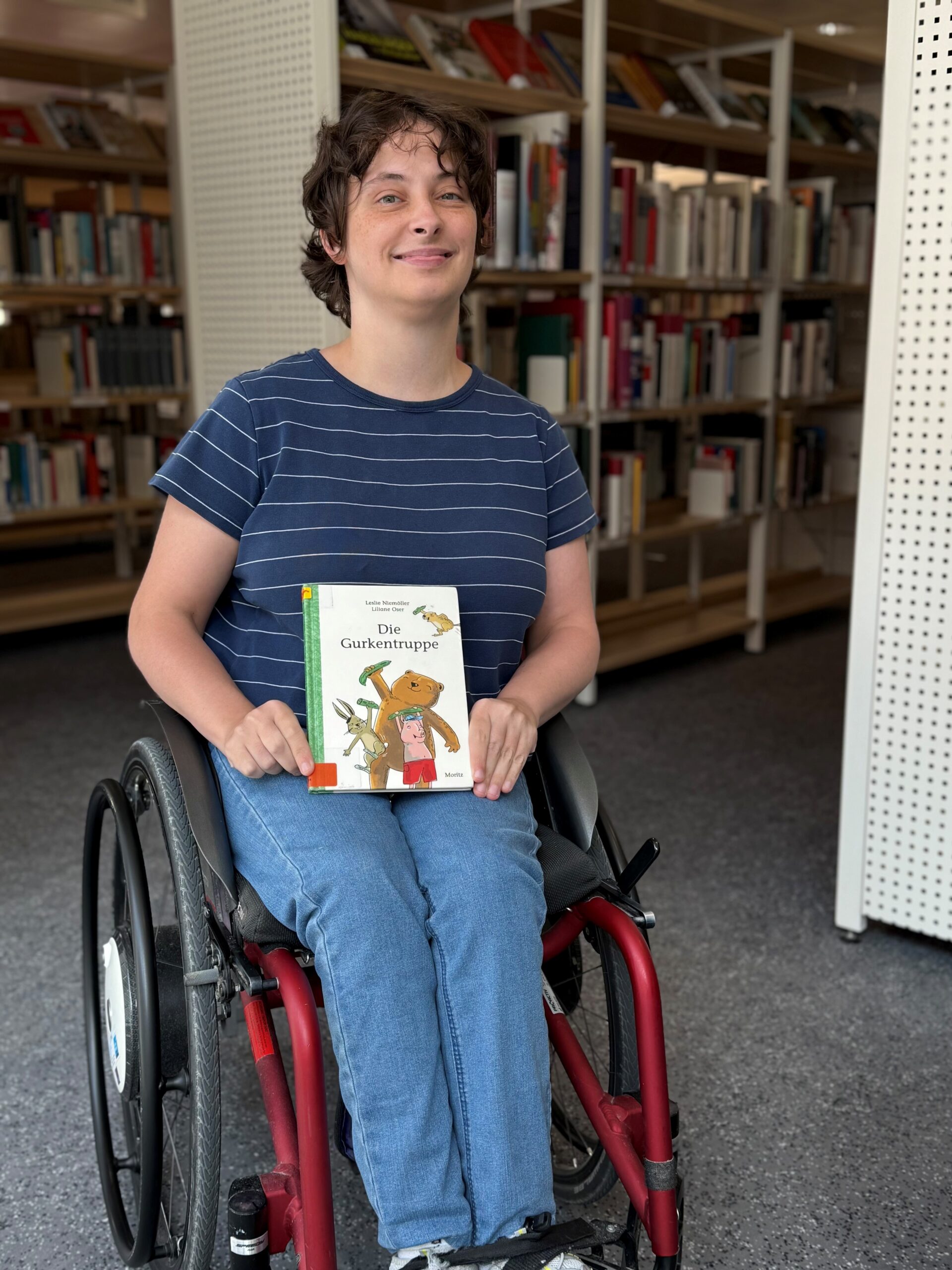 Eine junge Frau im Rollstuhl, die ein Kinderbuch in der Hand hält und in die Kamera lächelt.