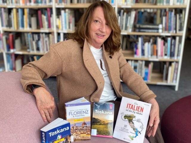 Eine Frau mit mittellangen braunen Haaren in einem Mantel sitzt auf einem rosa Sessel. Vor ihr stehen vier Bücher.