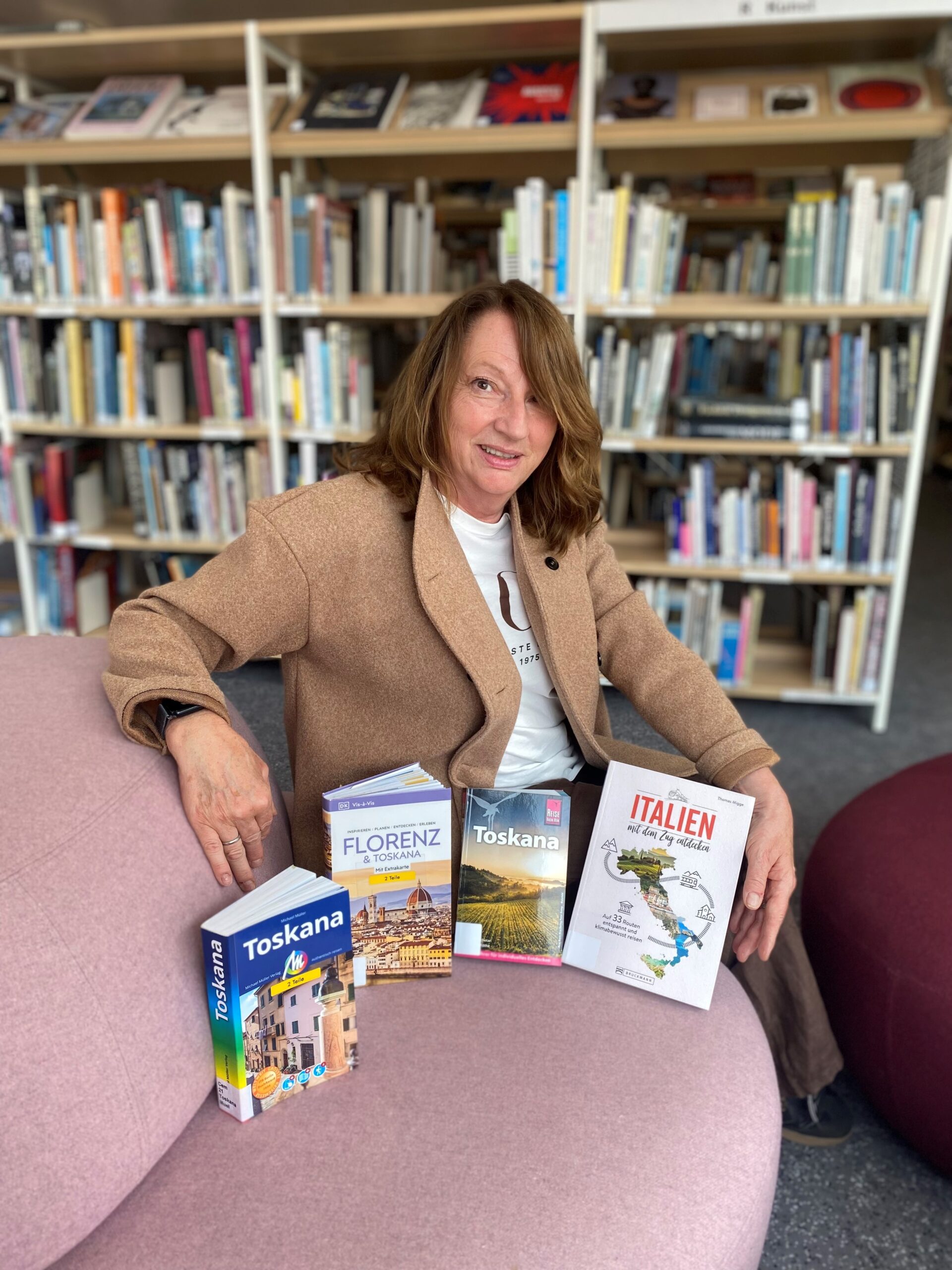Eine Frau mit mittellangen braunen Haaren in einem Mantel sitzt auf einem rosa Sessel. Vor ihr stehen vier Bücher.