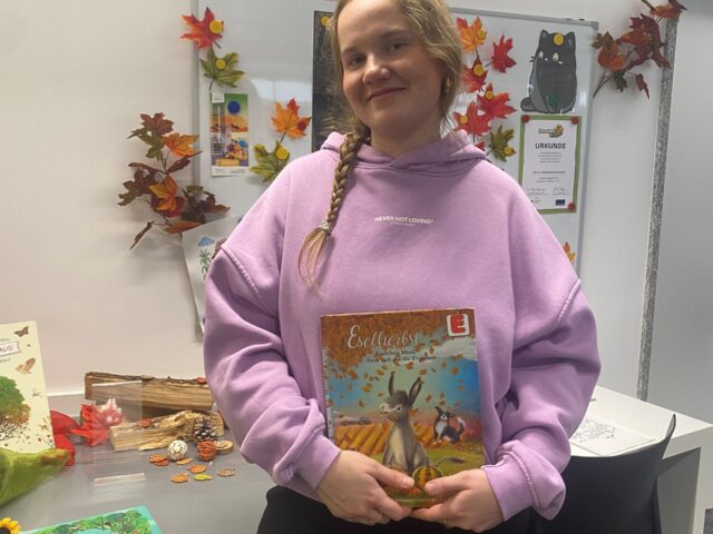 Eine junge Frau mit langen blonden Haaren und in einem rosa Pullover hält ein Bilderbuch in der Hand und lächelt in die Kamera.