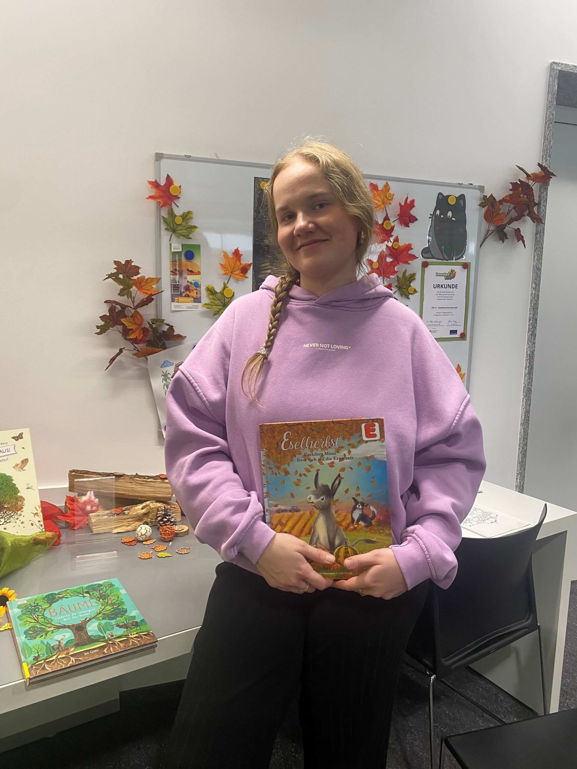 Eine junge Frau mit langen blonden Haaren und in einem rosa Pullover hält ein Bilderbuch in der Hand und lächelt in die Kamera.