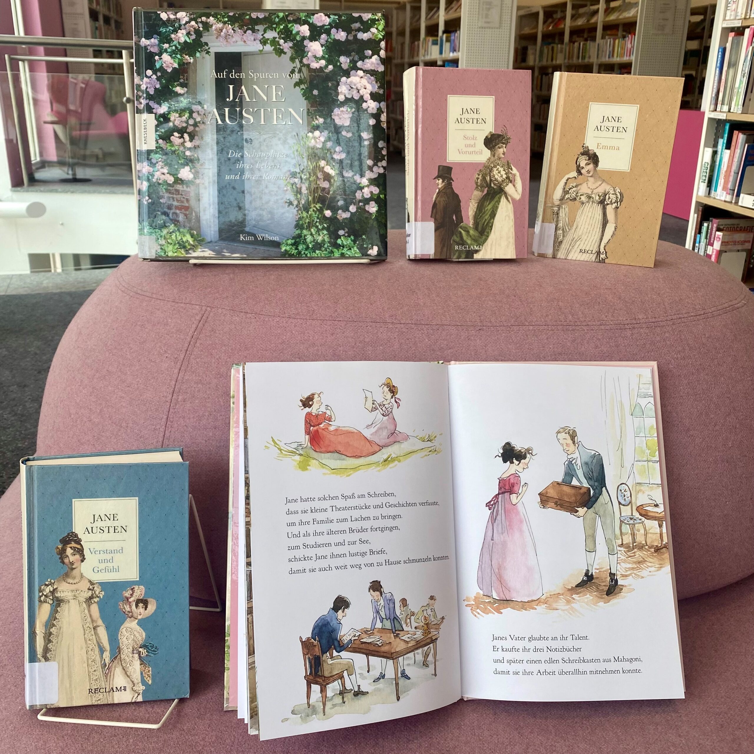Eine Reihe von Büchern über und von Jane Austen, die auf einer rosanen Couch präsentiert werden.