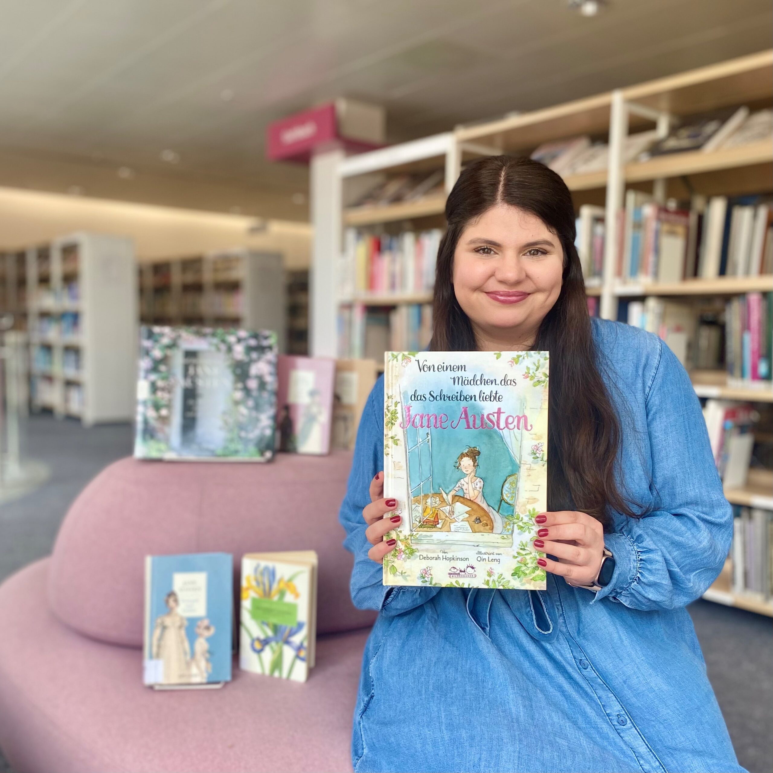 Eine junge Frau mit langen dunklen Haaren und einem blauen Kleid, hält lächelns ein Buch in die Kamera. Sie sitzt auf einem Sofa, im Hintergrund sind Bücherregale zu sehen.