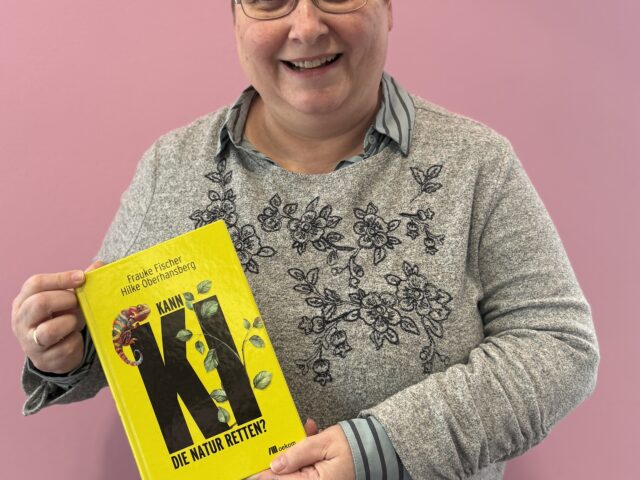 Eine Frau mit kurzen Haaren und Brille hält ein gelbes Buch in den Händen.