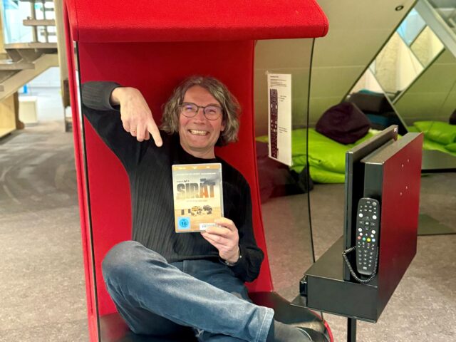 Ein Mann sitzt grinsend in einem roten Filmsessel und zeigt auf einen Film in seiner Hand.