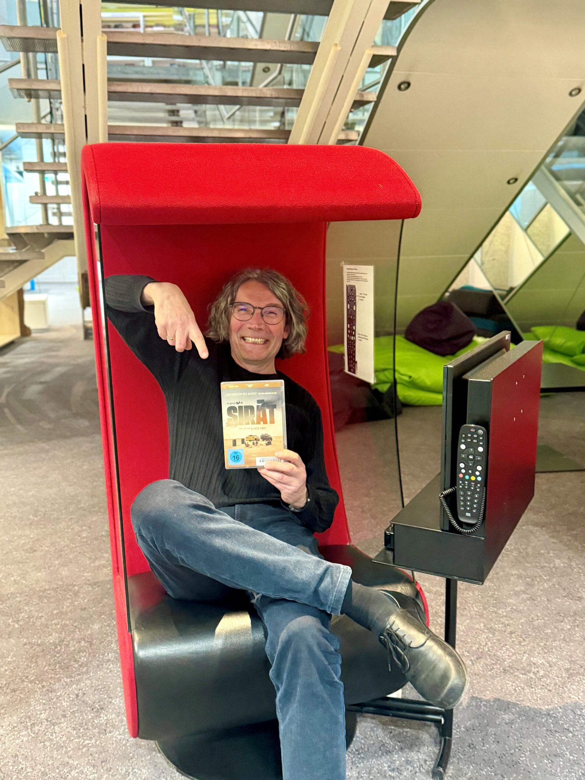 Ein Mann sitzt grinsend in einem roten Filmsessel und zeigt auf einen Film in seiner Hand.