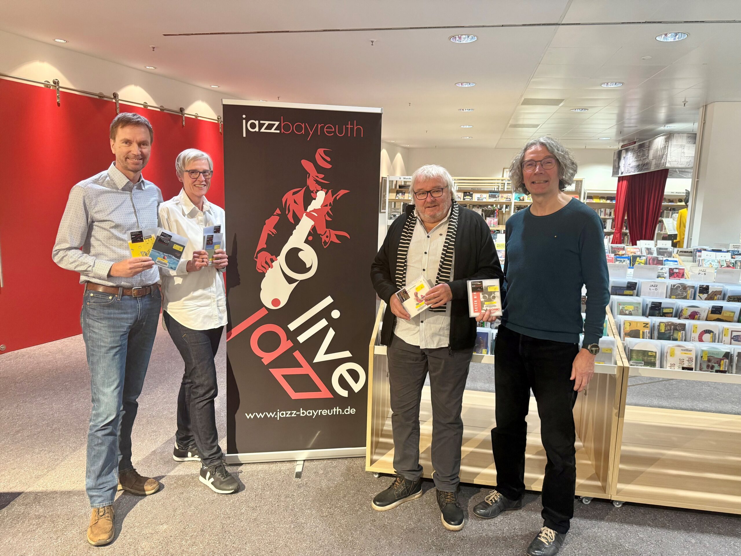 Vier Männer mit CDs in der Hand stehen neben einem Banner des Jazz-Vereins und lächeln.