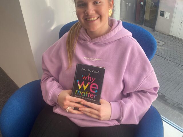 Eine junge Frau im rosa Pulli und mit zugebundenen Haaren sitzt in einem Sessel und hält ein Buch vor den Körper.