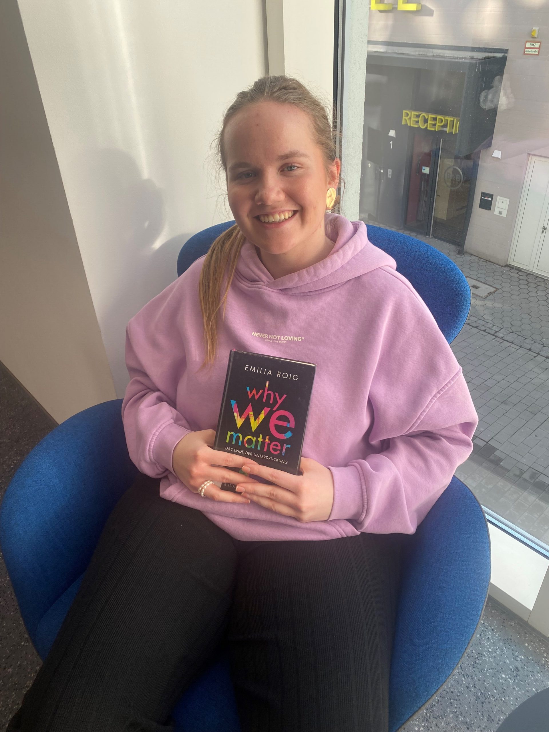 Eine junge Frau im rosa Pulli und mit zugebundenen Haaren sitzt in einem Sessel und hält ein Buch vor den Körper.