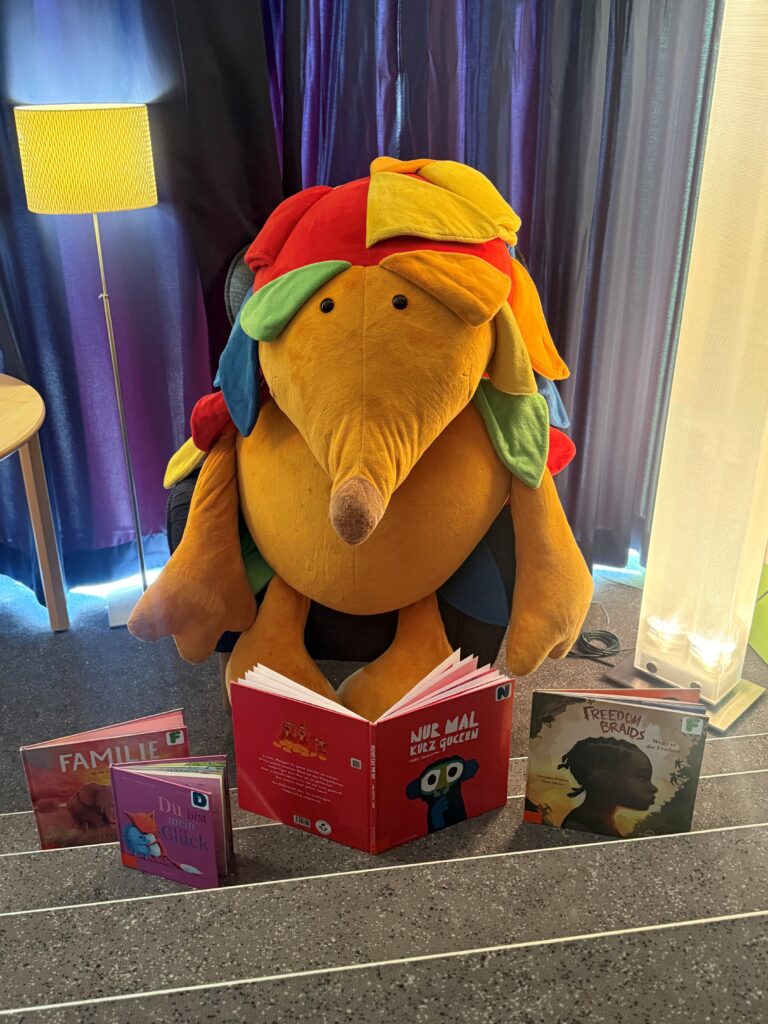 Ein großer Plüsch-Igel, der in einem Sessel sitzt. Vor ihm sind einige Bilderbücher.