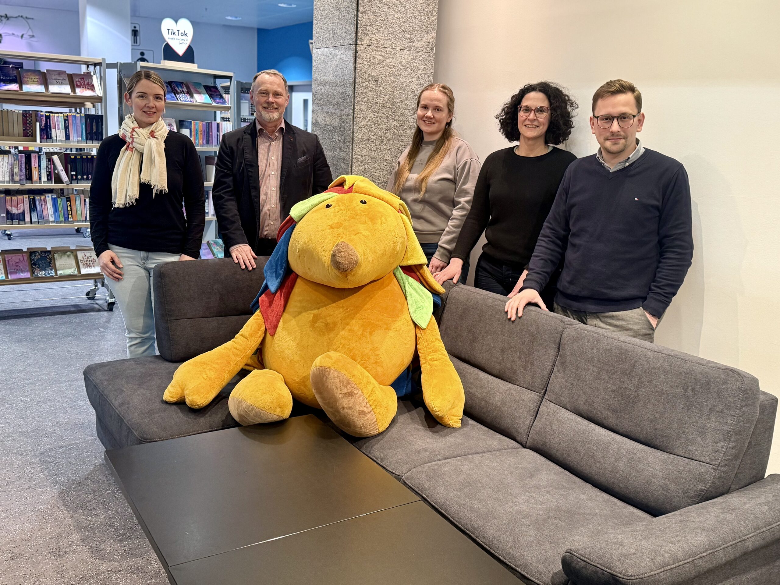 Drei Frauen und zwei Männer stehen hinter einem Sofa, auf dem ein großes Kuscheltier sitzt