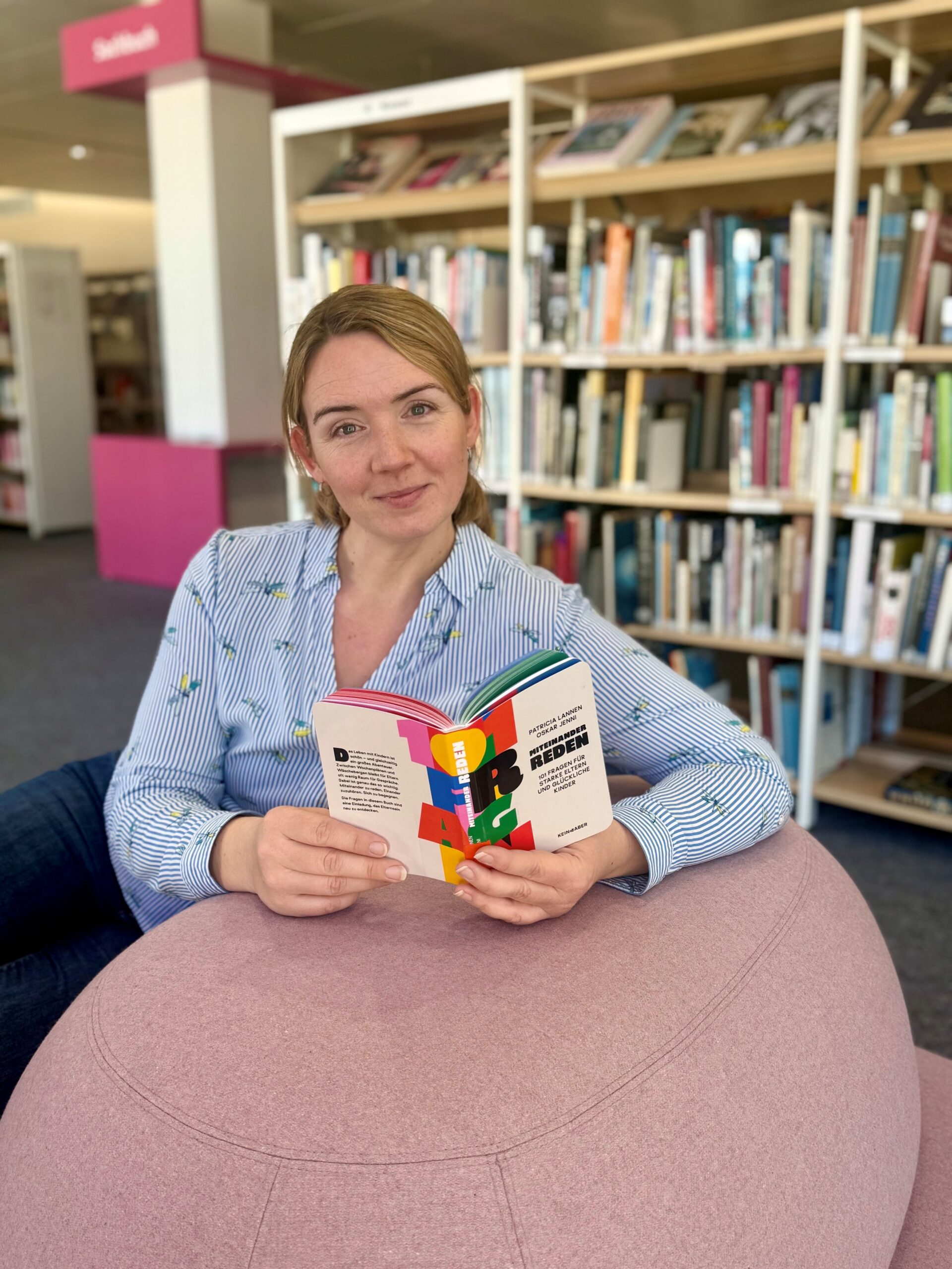 Eine junge Frau sitzt auf rosa Sessel mit einem Buch in der Hand und lächelt in die Kamera.