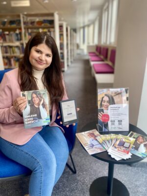 Eine junge Frau mit langen braunen Haaren und einem rosa Blazer sitzt auf einem Sessel und hält ein eBook-Reader und eine Broschüre in der Hand. Auf dem Tisch neben ihr sind einige Flyer ausgelegt.