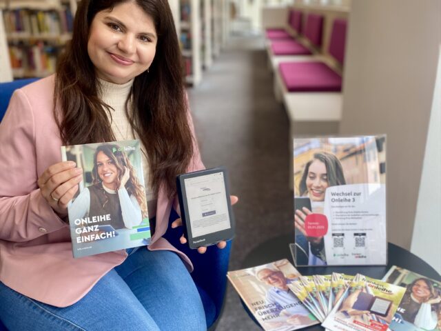Eine junge Frau mit langen braunen Haaren und einem rosa Blazer sitzt auf einem Sessel und hält ein eBook-Reader und eine Broschüre in der Hand. Auf dem Tisch neben ihr sind einige Flyer ausgelegt.