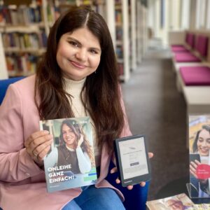 Eine junge Frau mit langen braunen Haaren und einem rosa Blazer sitzt auf einem Sessel und hält ein eBook-Reader und eine Broschüre in der Hand. Auf dem Tisch neben ihr sind einige Flyer ausgelegt.
