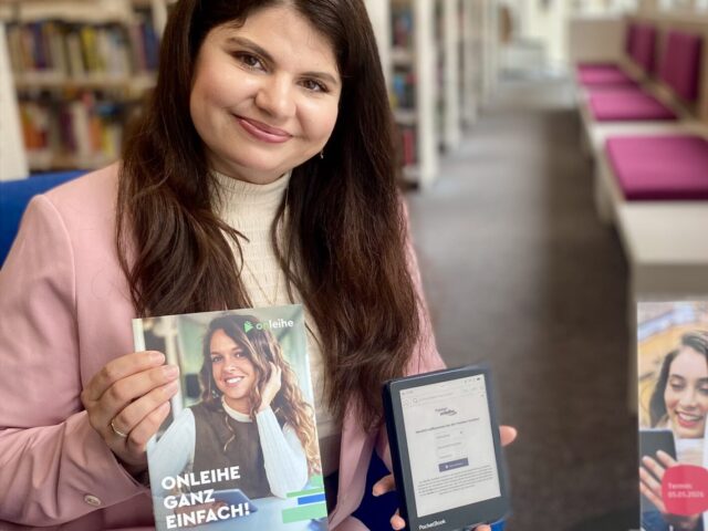 Eine junge Frau mit langen braunen Haaren und einem rosa Blazer sitzt auf einem Sessel und hält ein eBook-Reader und eine Broschüre in der Hand. Auf dem Tisch neben ihr sind einige Flyer ausgelegt.